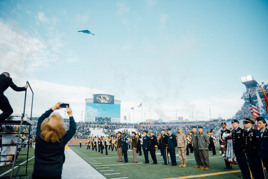 A photo of a Northrup Grumman B-2 Spirit flying over Faurot Field and assembled representatives of the U.S. Armed Forces during Military Appreciation Day.